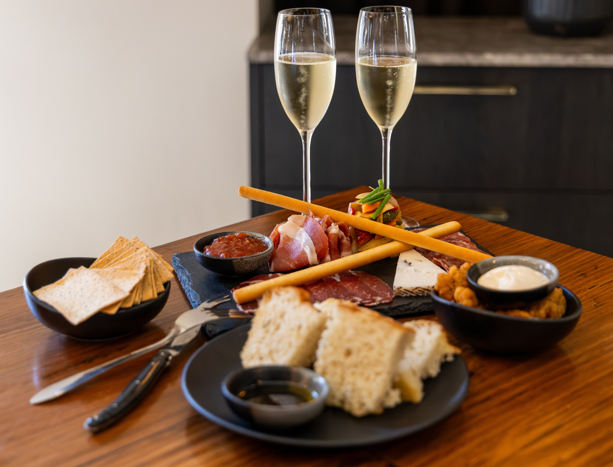 A charcuterie board spread on a wooden table with two glasses of white wine, sliced bread, crackers, small bowls of dips and spreads, and a dark plate in a real indoor setting.