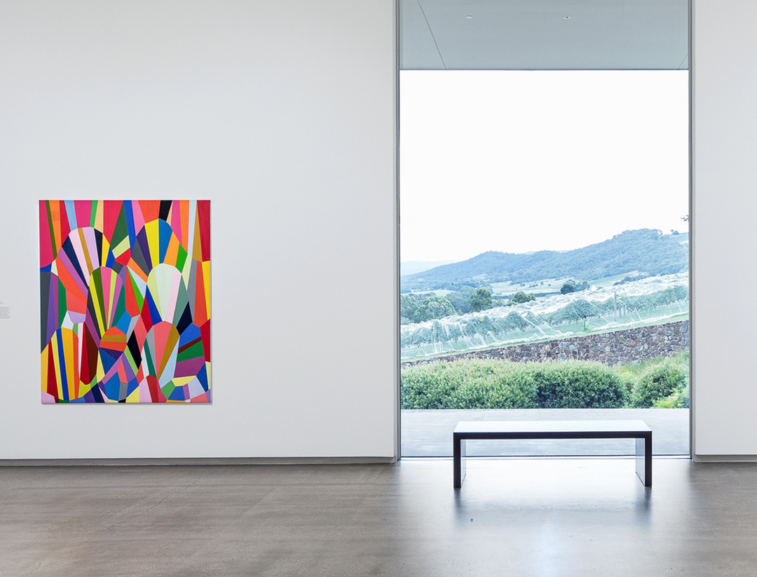 A colorful abstract geometric painting hangs on a white gallery wall next to a large window showing a landscape view, with a black bench on a polished floor below.