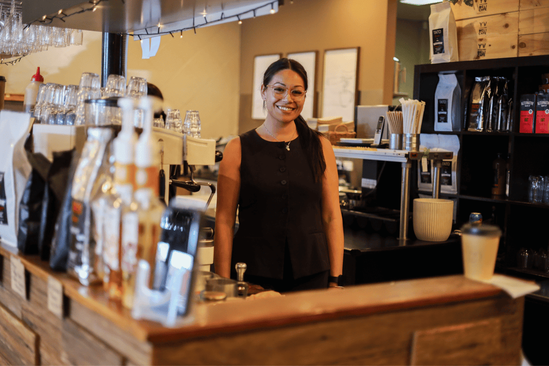 A smiling woman with glasses and a ponytail stands behind a wooden coffee bar counter in a cafe setting, wearing a black sleeveless top.