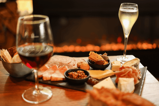 A styled charcuterie board with cheeses, crackers, and small dishes on a wooden table, with two wine glasses in the foreground and a warm fireplace blurred in the background.