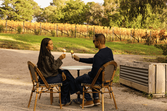 A man and a woman sit at a small outdoor table in a vineyard setting, each holding an ice cream cone and clinking them together. The scene includes wicker chairs, a gravel path, and rows of grapevines in the background.