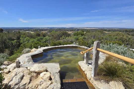 A natural stone outdoor hot spring or pool overlooking a vast landscape of trees and hills under a clear blue sky, with a wooden handrail along the edge.