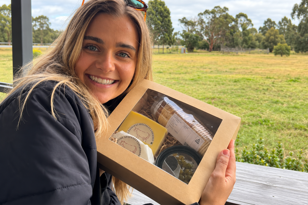 A smiling woman outdoors holds a kraft gift box with a clear window showing packaged items inside, with a grassy field and trees in the background.