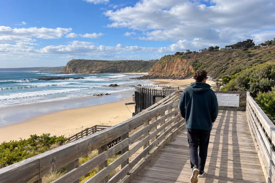 A person in a dark hoodie and black pants walks along a wooden boardwalk overlooking a sandy beach, ocean waves, and rocky cliffs under a blue sky with white clouds.