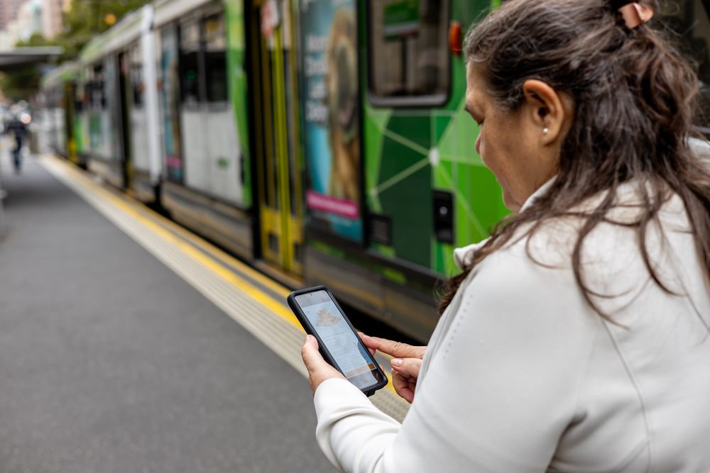A woman in a white jacket stands on a train platform holding a smartphone, with a green and yellow city train passing in the background.