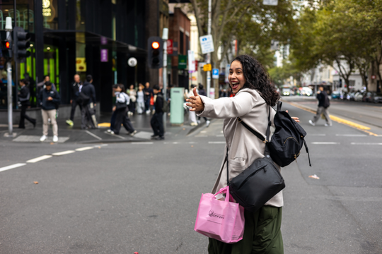 A woman standing on a city street crosswalk wearing a light beige jacket, dark green pants, and carrying a black backpack and a bright pink tote bag. She is smiling and gesturing with her hand.
