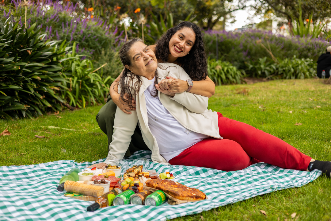 Two women hugging while sitting on a green and white checkered picnic blanket on a grassy lawn, with a picnic spread of food in front of them and garden plants in the background.