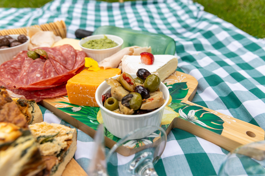A picnic-style spread of assorted cheeses, cured meats, olives, crackers, and dips arranged on a green-and-white checkered blanket outdoors, with grass visible in the background.