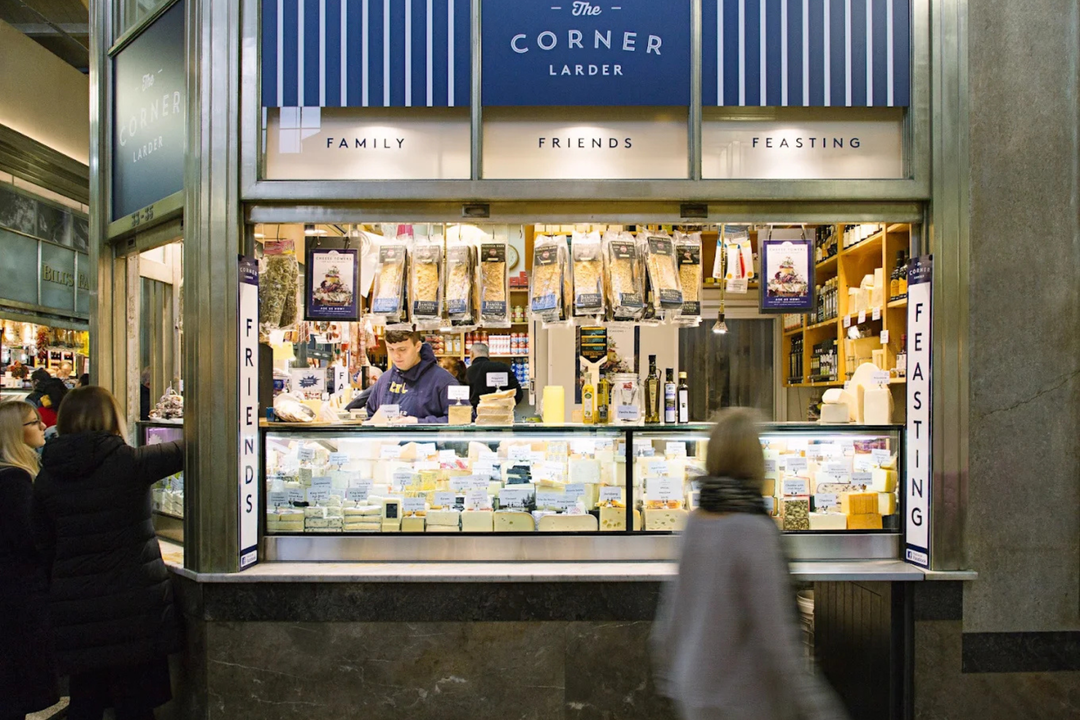 The Corner Larder retail shop front with a blue and white striped sign, displaying an array of packaged meats, cheeses, and other grocery items behind a glass counter. Customers are visible walking past the storefront.