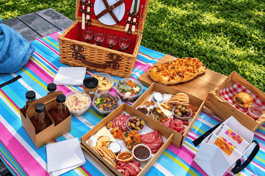 A colorful outdoor picnic spread on a striped blanket with a wicker picnic basket, charcuterie boards, salads, drinks, and a pizza on a grassy lawn.