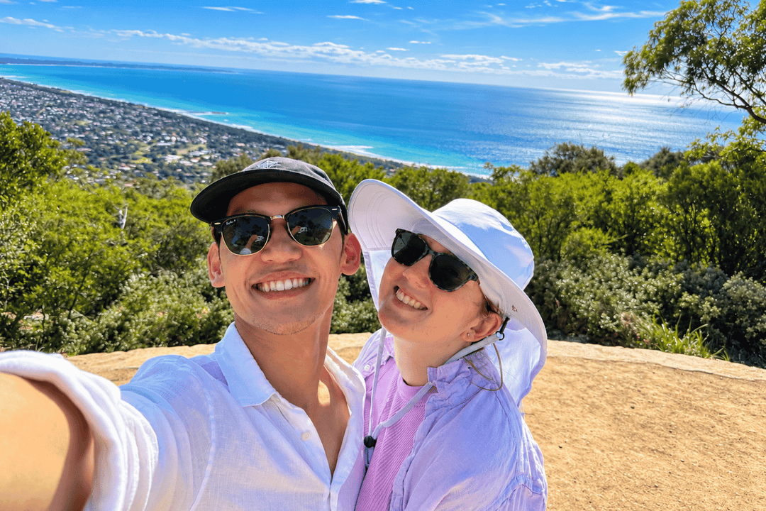 A smiling couple taking a selfie outdoors on a hilltop with a scenic ocean and coastline in the background. The man wears a white polo shirt, black cap, and sunglasses, while the woman wears a white wide-brim hat, sunglasses, and a light pink shirt.