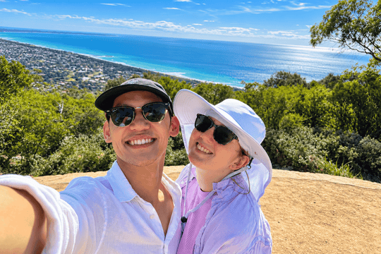 A smiling couple taking a selfie outdoors on a hilltop with a scenic ocean and coastline in the background. The man wears a white polo shirt, black cap, and sunglasses, while the woman wears a white wide-brim hat, sunglasses, and a light pink shirt.