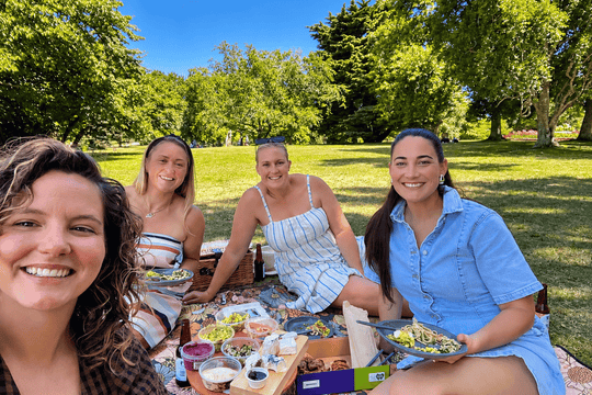 Four women smiling and posing outdoors on a picnic blanket in a sunny park with green grass and trees. Plates of food and a picnic setup are spread on the blanket in front of them.