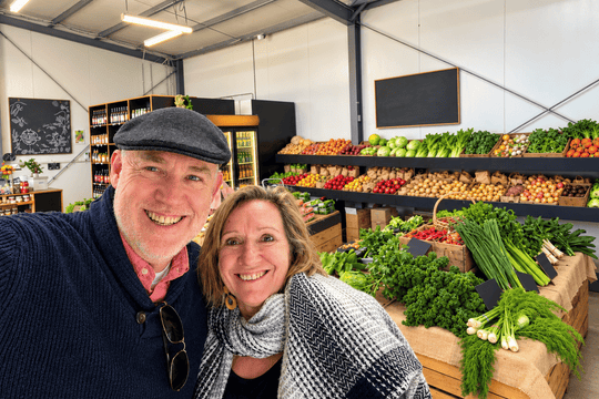 A smiling man and woman pose for a selfie inside a farm-to-table grocery store or market, with fresh vegetables and fruits displayed on shelves in the background.