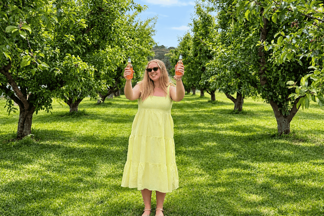 A woman wearing a light yellow tiered midi dress and sunglasses stands in a sunny apple orchard, holding up two bottles of water.