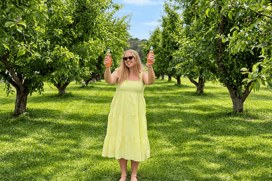 A woman wearing a light yellow tiered midi dress and sunglasses stands in a sunny apple orchard, holding up two bottles of water.