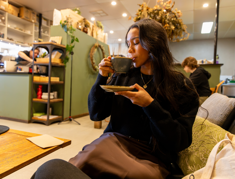 A woman with long dark hair sits on a couch in a cozy cafe setting, holding a dark ceramic mug and saucer. She is wearing a black knit sweater and brown pants.