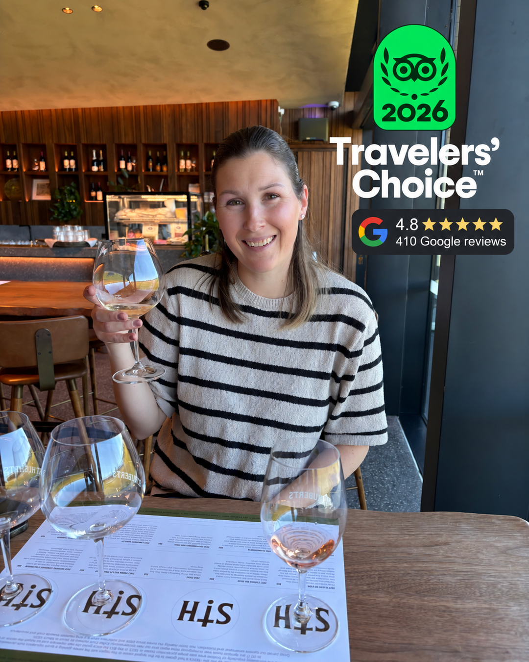 A woman smiling at a restaurant table holding a wine glass, with multiple wine glasses on the table and a 'Travelers' Choice 2026' badge overlay in the top right corner.