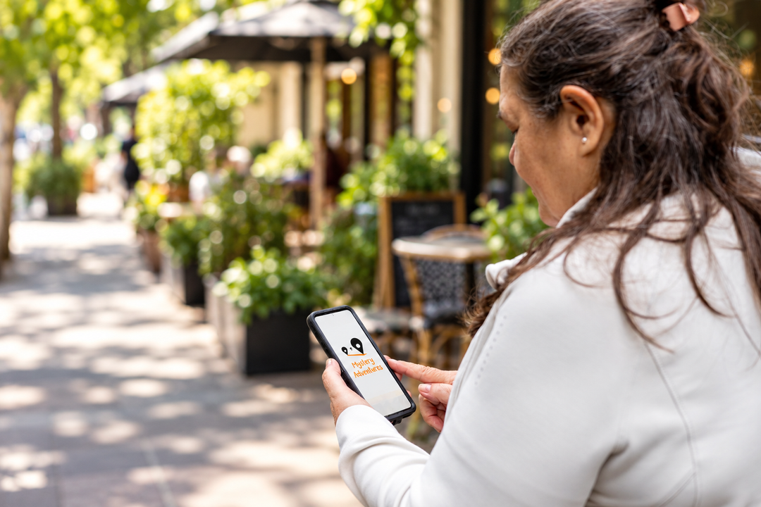 A woman in a white jacket stands on a sunlit outdoor cafe walkway, holding a smartphone that displays a delivery app interface.